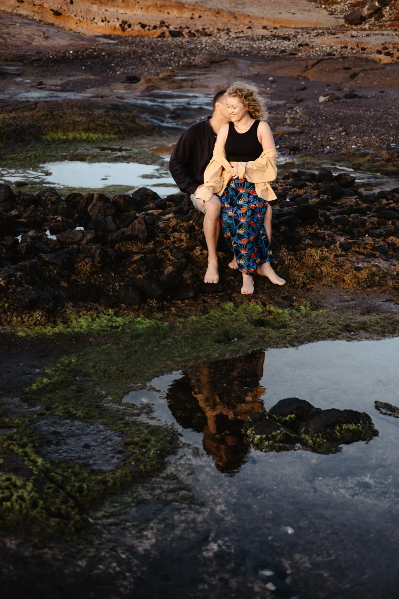 Pareja romántica sentada rocas con serenos reflejos agua durante puesta sol