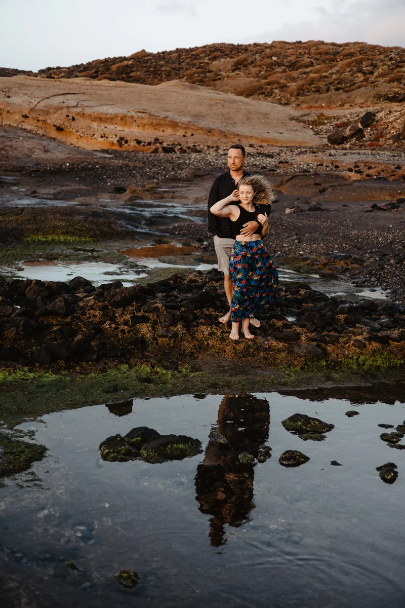 Tierno abrazo pareja contra dramático paisaje volcánico Tenerife con reflejos piscinas marea