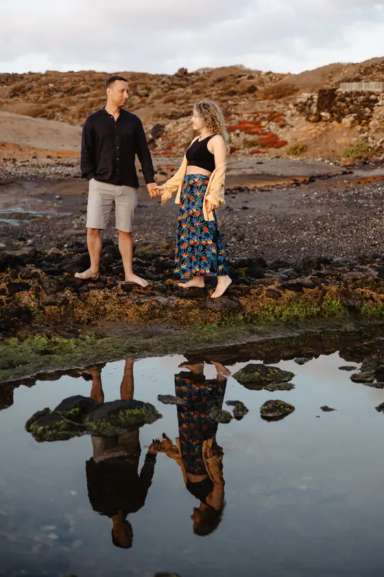 Walking couple holding hands on black volcanic rocks with perfect reflections