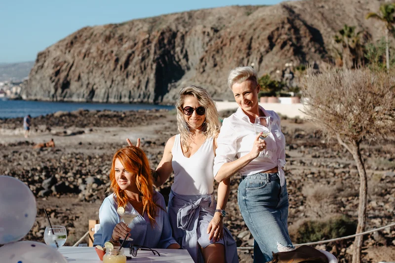 Three joyful women with balloons against volcanic cliffs and ocean backdrop in Tenerife