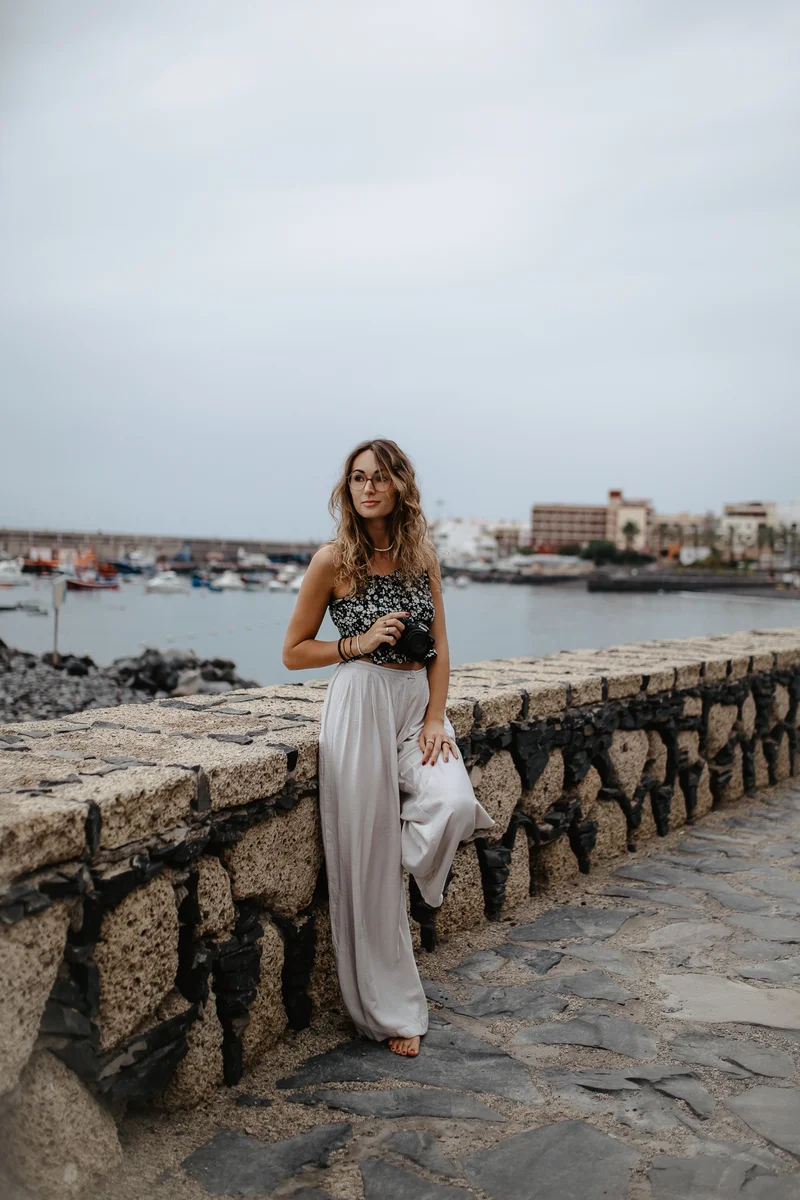 Mujer posando con cámara en paseo marítimo de piedra con barcos de fondo en Tenerife