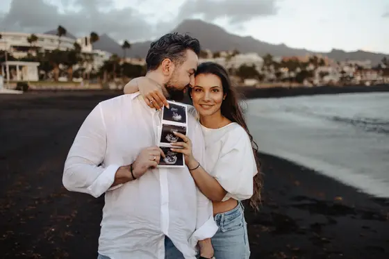 Happy couple posing with ultrasound photos during pregnancy session on volcanic beach