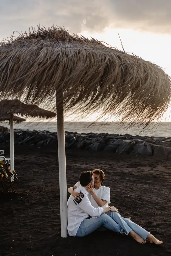 Relaxed sitting pose of couple under palm umbrella on black volcanic beach