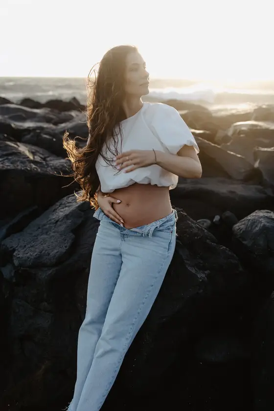 Tender pose of pregnant woman touching belly on volcanic rocks in golden light