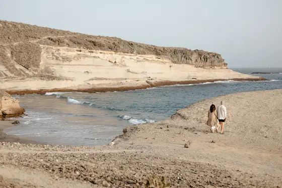 Pareja caminando por la playa salvaje de La Caleta con acantilados volcánicos dramáticos