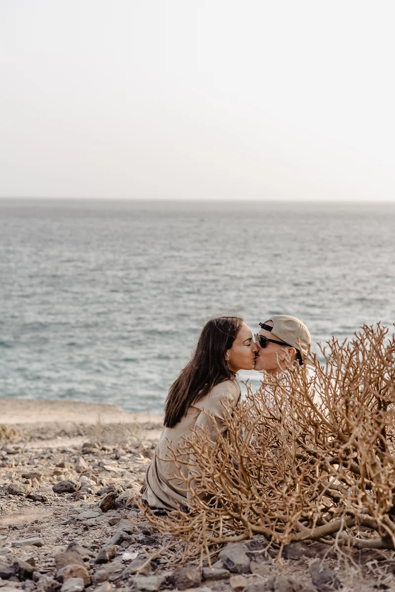Pareja besándose románticamente entre plantas con telón de océano en La Caleta