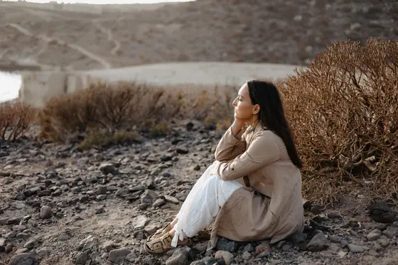 Mujer en vestido beige sentada contemplativamente entre vegetación seca en acantilados La Caleta