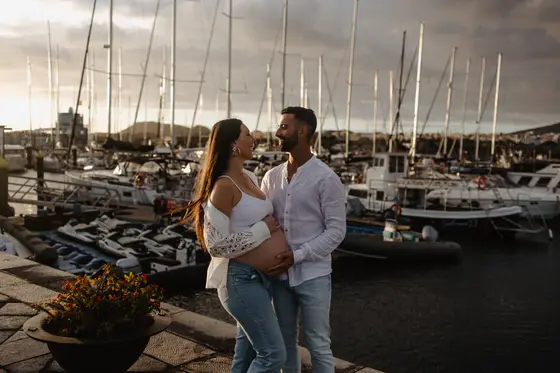 Pareja esperando bebé abrazándose en el puerto de Las Galletas con veleros de fondo durante la hora dorada