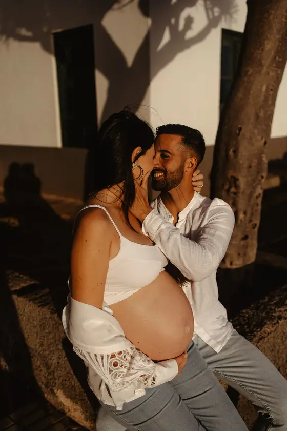 Pareja tierna esperando bebé en abrazo romántico bajo palmera durante sesión fotográfica familiar en Las Galletas
