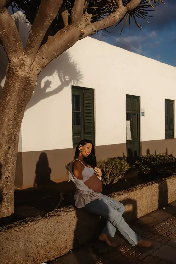Mujer embarazada feliz sentada junto a arquitectura canaria bajo palmera en luz dorada durante sesión de maternidad
