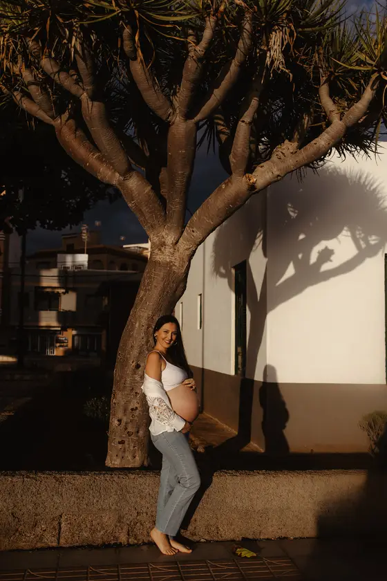 Futura madre sonriente en vestido blanco posando junto a casa canaria bajo palmera durante la hora dorada