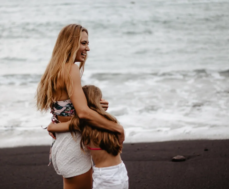 Happy mother hugging daughter on dark beach with white ocean foam backdrop in Tenerife photography