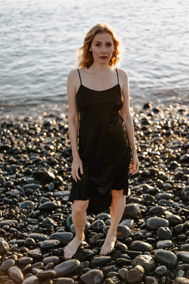 Elegancka blondynka w czarnej sukni na skalistej plaży Teneryfy podczas złotej godziny