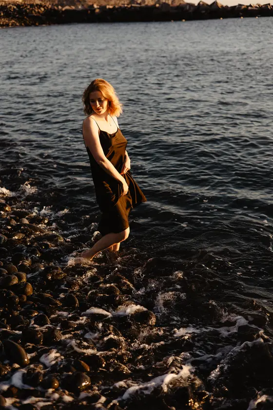 Smiling woman standing in shallow ocean water on rocky Tenerife beach during golden sunset hour