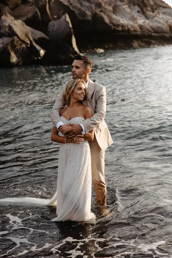 Newlyweds embracing while standing in shallow ocean water against volcanic rocks at Playa Paraiso