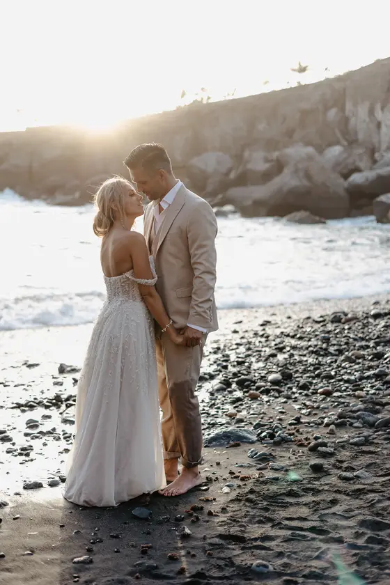 Newlyweds standing barefoot on black volcanic beach in golden sunlight at Playa Paraiso