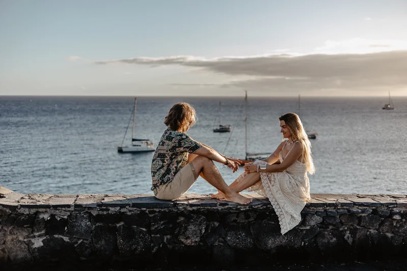 Pareja sentada en muro de piedra con vista a yates y aguas océano Atlántico