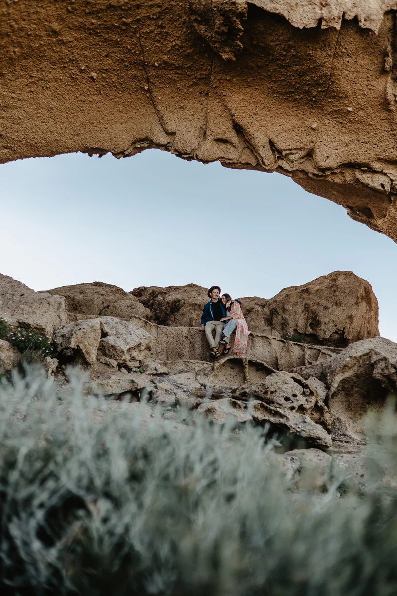 Couple sitting on rocks under arch with view of desert landscape and succulent vegetation