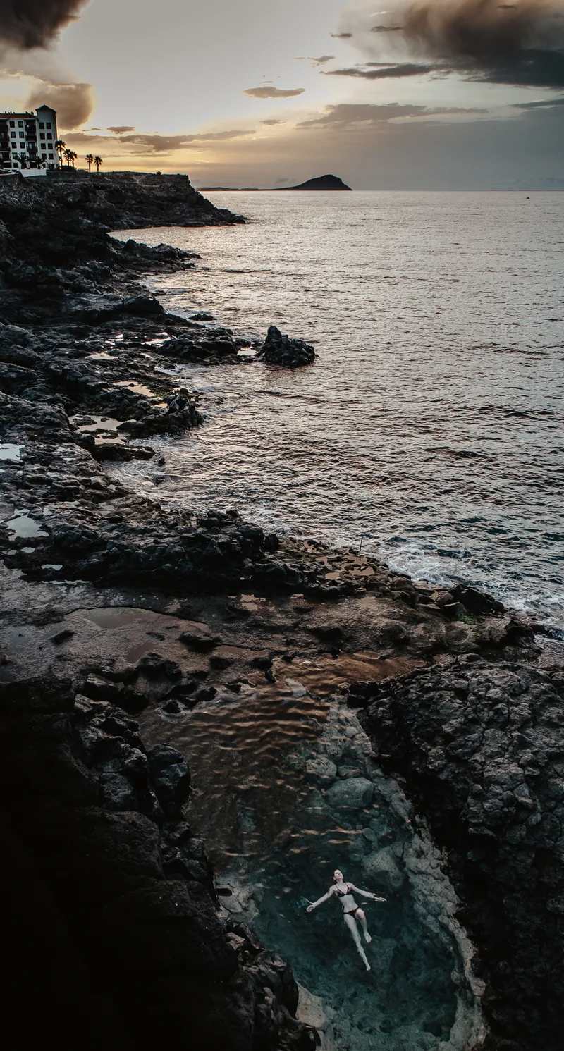 Mujer nadando en una piscina natural entre rocas volcánicas negras con cielo dramático en Tenerife