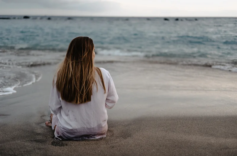 Kobieta w ciąży siedzi tyłem na piaszczystej plaży patrząc na ocean