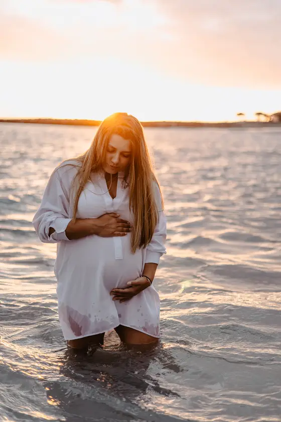 Intimate portrait of pregnant woman holding belly in ocean at sunset