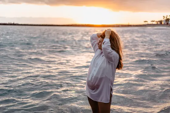 Pregnant woman with arms raised standing in ocean during sunset