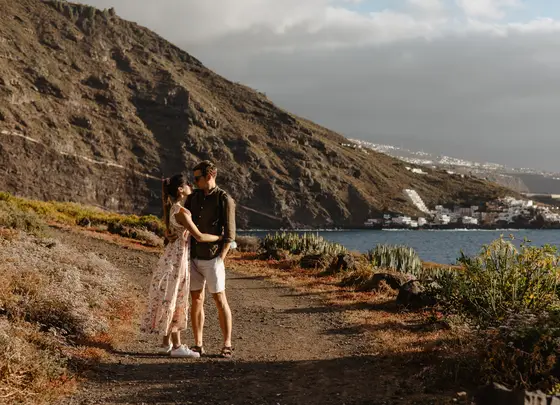 Pareja abrazándose en costa volcánica de Tenerife con vista al océano y casas blancas costeras