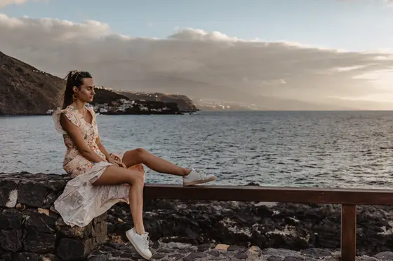 Mujer en vestido blanco sentada en plataforma de madera en la costa de Tenerife durante el atardecer