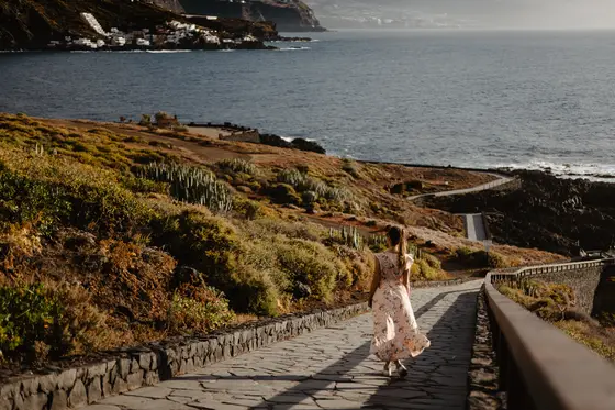 Mujer en vestido caminando sendero de piedra sobre océano en costa de Tenerife durante hora dorada