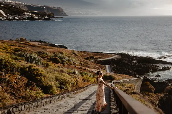 Mujer en vestido caminando sendero costero de piedra a lo largo de la costa de Tenerife con vistas al océano