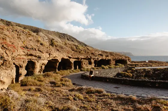 Cuevas ancestrales talladas en rocas costeras de Tenerife con mujer caminando sendero de piedra