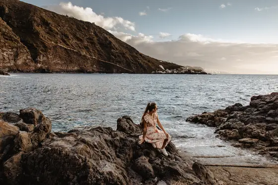 Mujer en vestido en rocas volcánicas junto al océano en la costa de Tenerife con vistas a acantilados