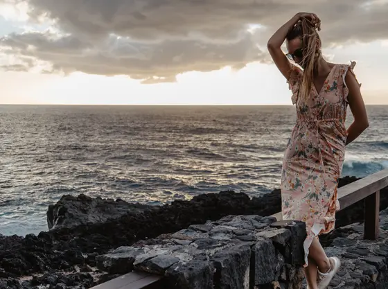 Mujer en vestido con patrón floral de pie en rocas volcánicas sobre el océano en Tenerife