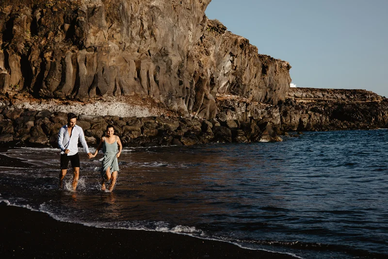 Pareja española enamorada caminando en olas del océano con acantilados de Tenerife de fondo