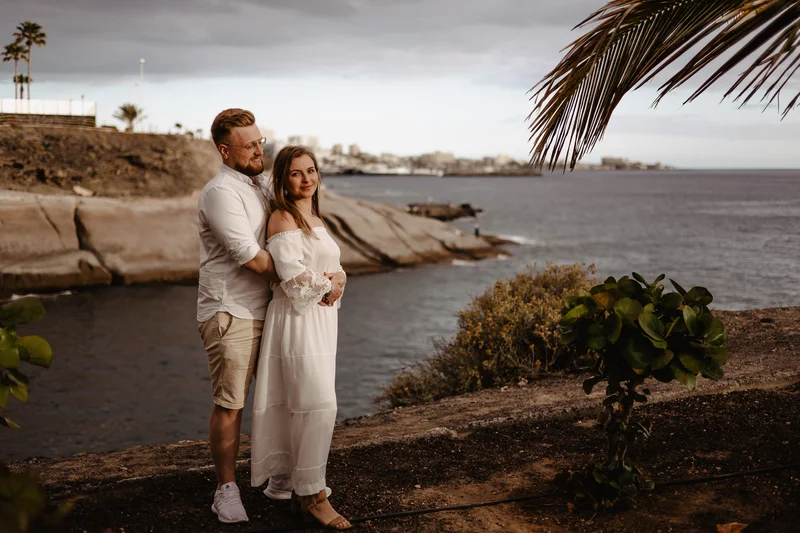 Pareja comprometida abrazada en costa de Tenerife con océano y palmeras en escenario romántico