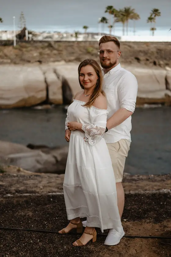 Elegante pareja comprometida en paseo marítimo de Tenerife con palmeras y océano de fondo