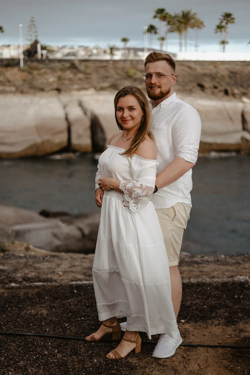 Elegante pareja comprometida en paseo marítimo de Tenerife con palmeras y océano de fondo