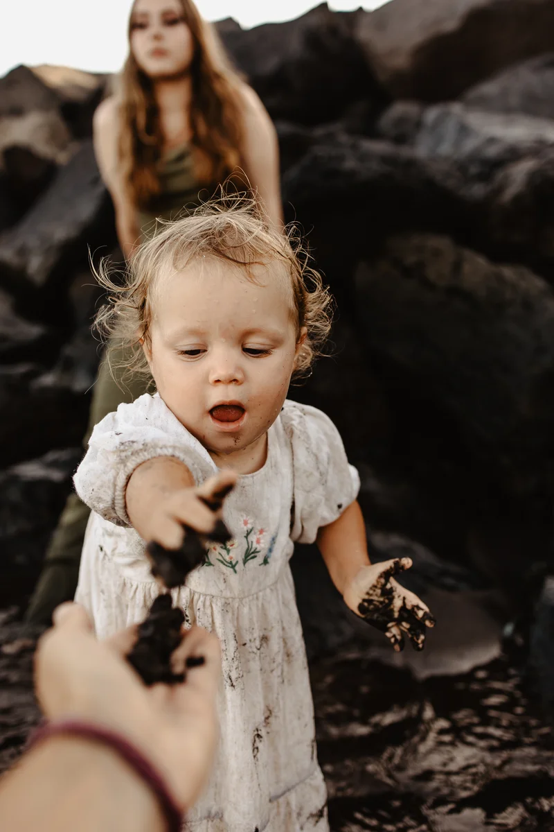 Małe dziecko wyciąga rączki podczas zabawy na plaży wulkanicznej Teneryfy z mamą w tle